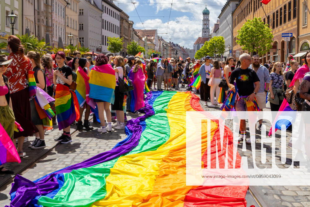 Teilnehmerinnen und Teilnehmer mit Regenbogenflaggen und bunten Kostümen nehmen am Christopher Street Day (CSD) in Augsburg teil. Der Demonstrationszug durch die Innenstadt wirbt für die Rechte von LGBTQ+-Menschen sowie für gesellschaftliche Vielfalt und Toleranz. Teilnehmer breiten eine große Regenbogenflagge, Regenbogen Fahne aus
