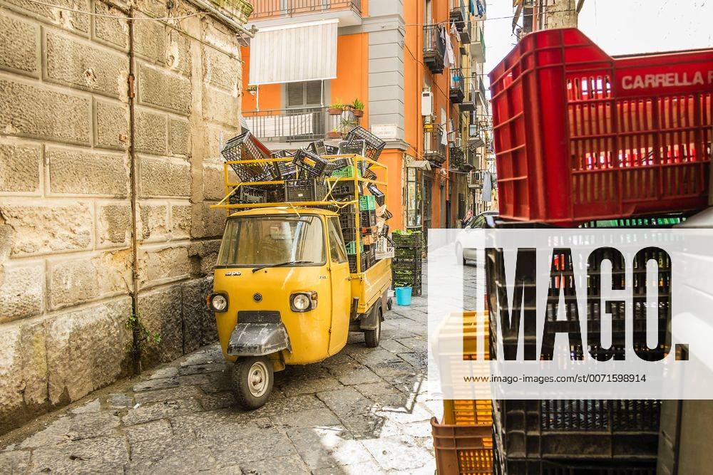 Eine Piaggio Ape mit aufgestapelten Kunstoffkisten steht in einer gepflasterten engen Gasse die