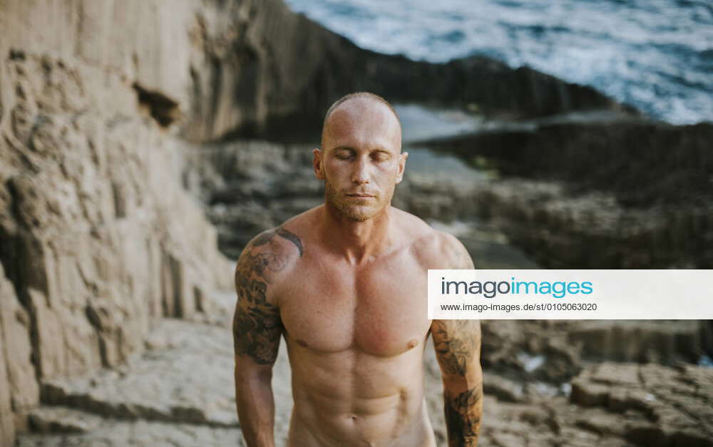 Tattooed nudist standing on volcanic rocks by the sea with closed eyes